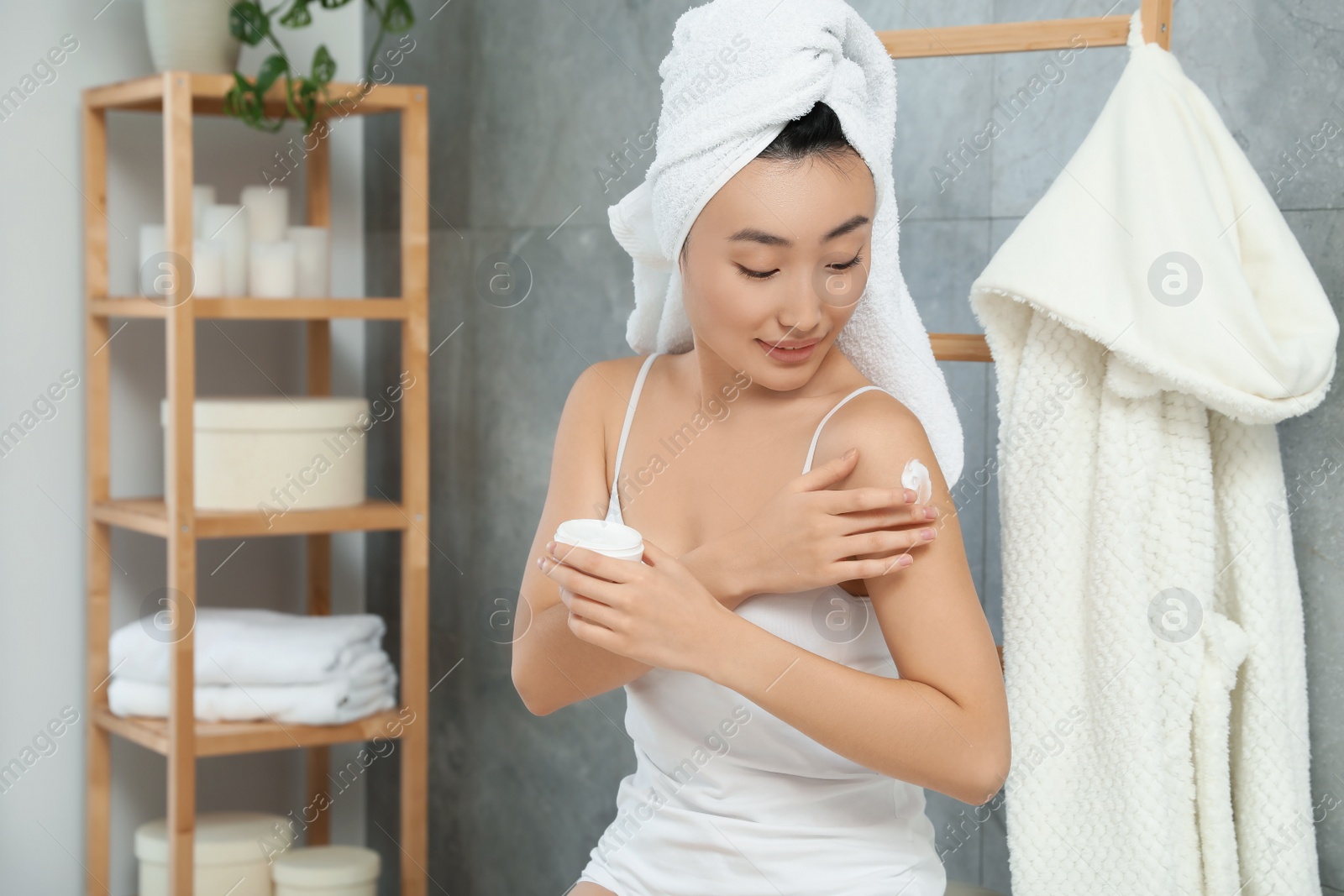 Photo of Beautiful young Asian woman applying body cream on shoulder in bathroom, space for text