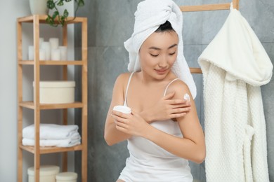 Photo of Beautiful young Asian woman applying body cream on shoulder in bathroom, space for text