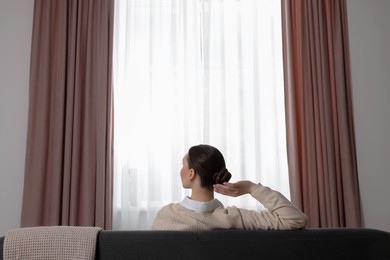 Photo of Woman sitting on sofa near window with stylish curtains at home, back view