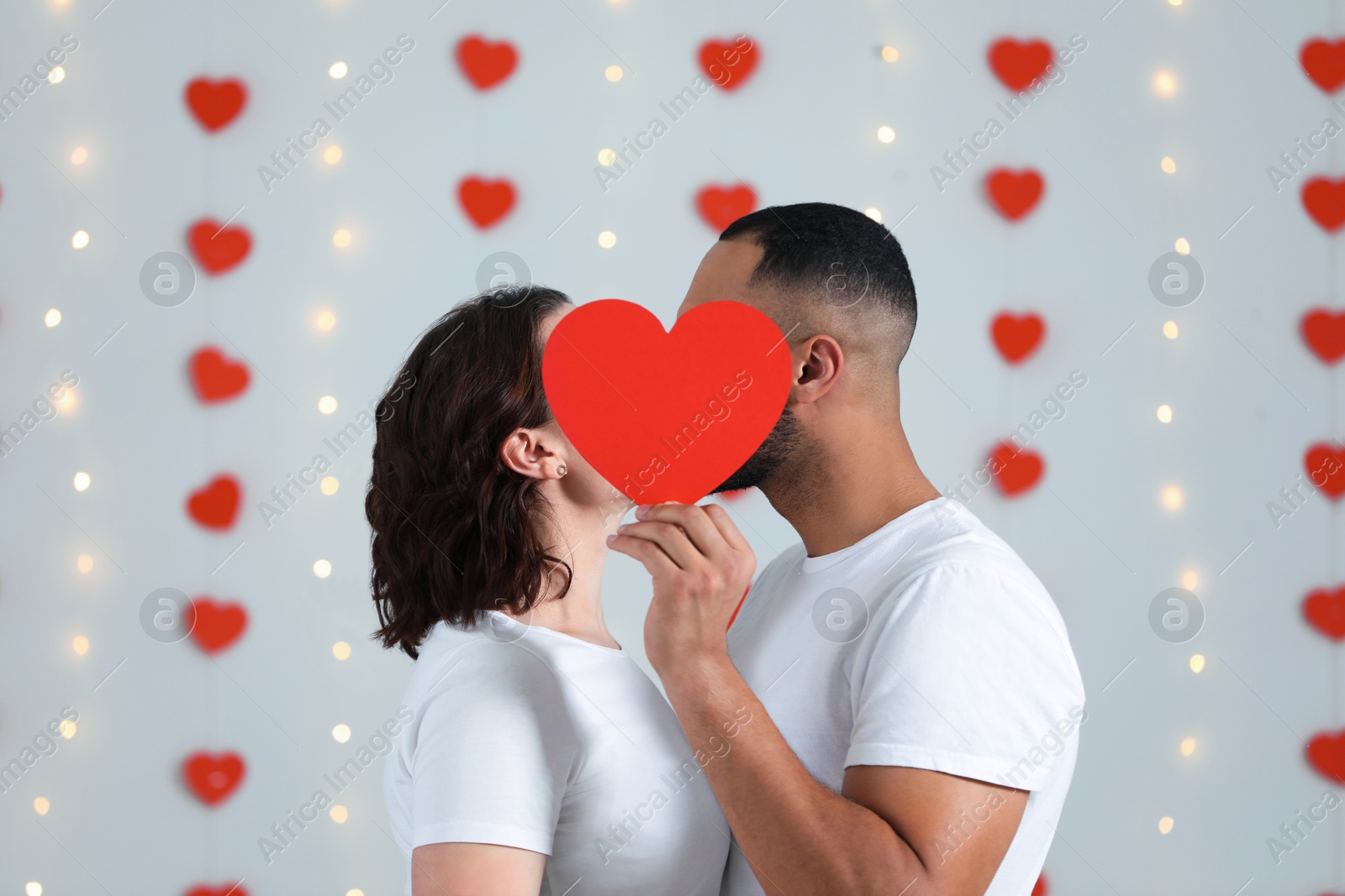 Photo of Lovely couple kissing behind red paper heart indoors. Valentine's day celebration