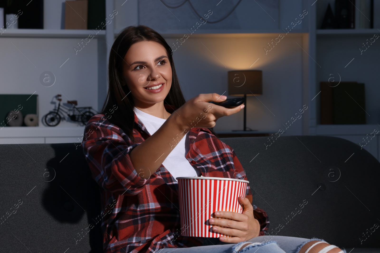Photo of Happy woman holding popcorn bucket and changing TV channels with remote control at home in evening