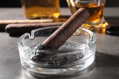 Photo of Ashtray with smoldering cigar and whiskey on grey table, closeup