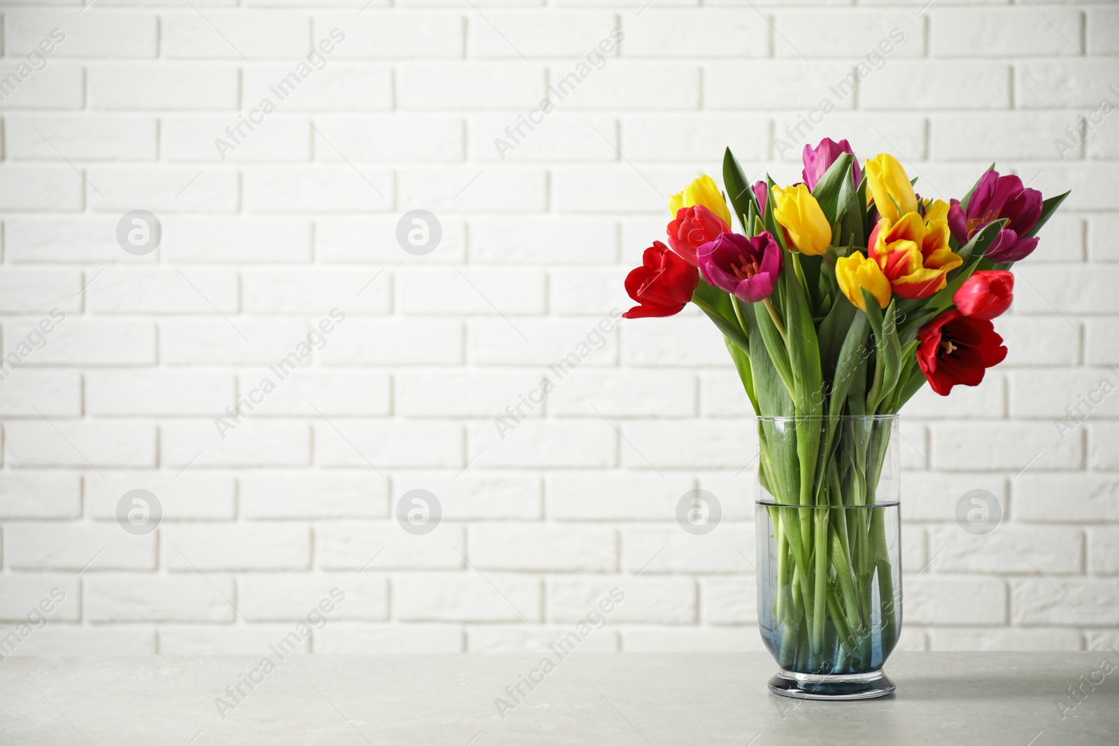 Photo of Beautiful spring tulips in vase on table near white brick wall. Space for text
