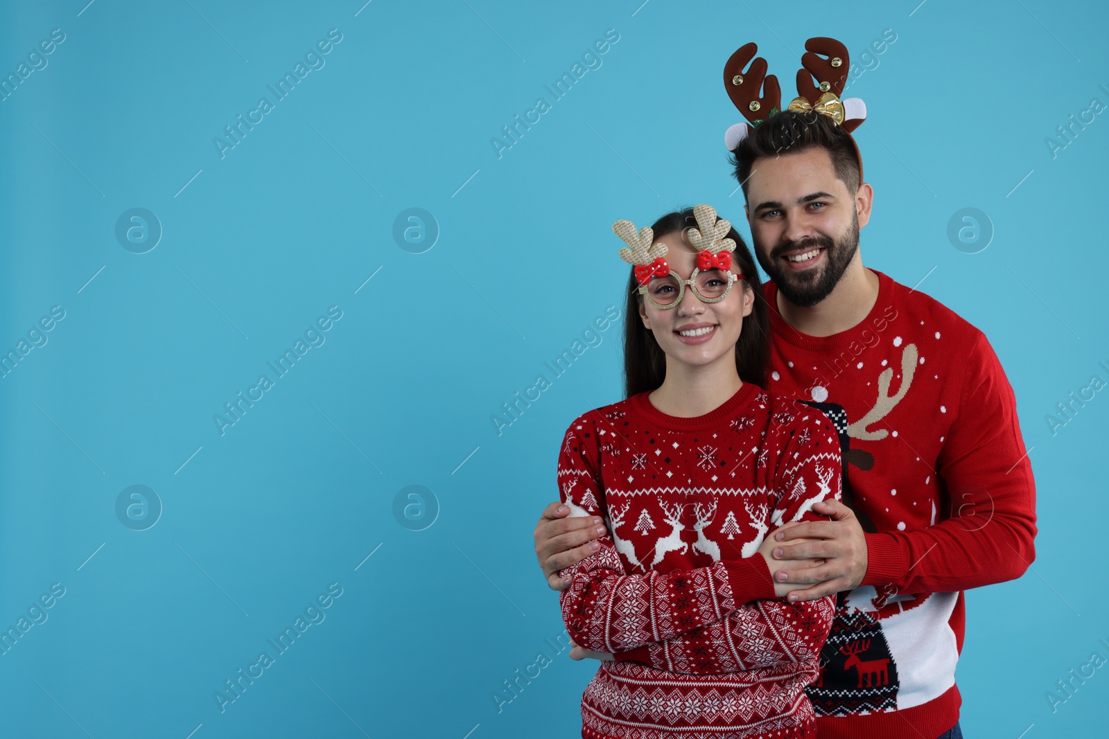 Photo of Happy young couple in Christmas sweaters, reindeer headband and funny glasses on light blue background. Space for text