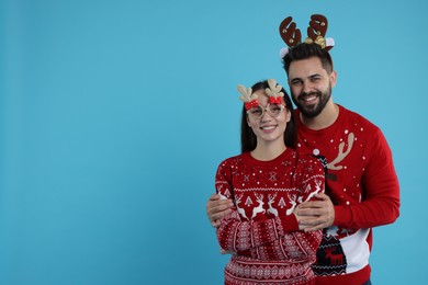 Photo of Happy young couple in Christmas sweaters, reindeer headband and funny glasses on light blue background. Space for text