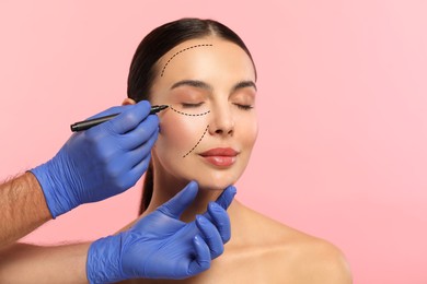 Woman preparing for cosmetic surgery, pink background. Doctor drawing markings on her face, closeup