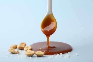 Photo of Spoon with tasty salted caramel, peanuts and salt on light blue background, closeup