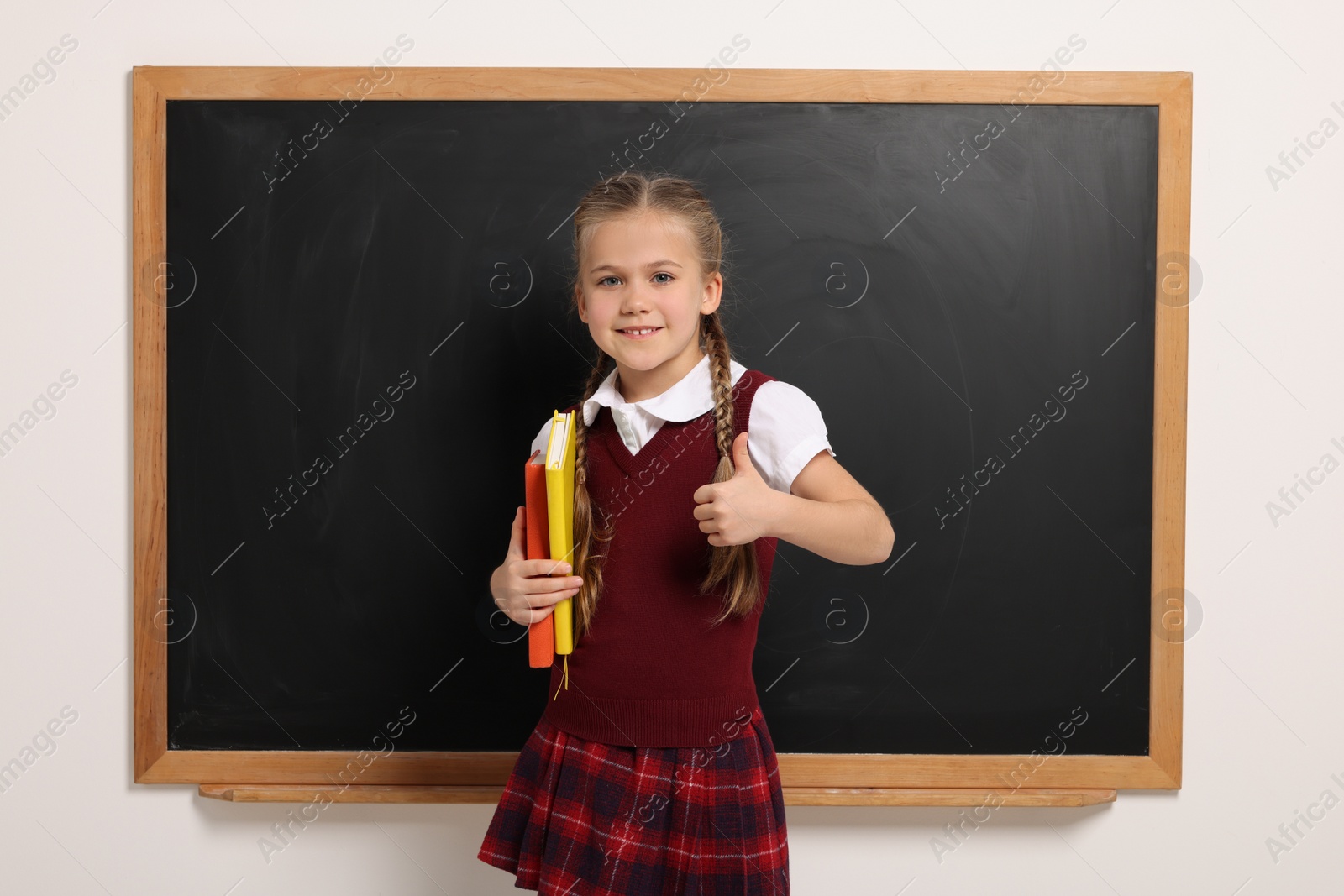 Photo of Happy schoolgirl with books showing thumb up gesture near blackboard