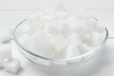 Photo of Many sugar cubes in glass bowl on white wooden table, closeup