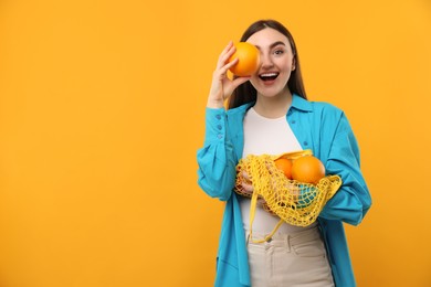 Woman holding string bag of fresh oranges and covering eye with fruit on orange background, space for text
