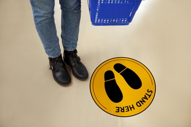Image of Keep social distance as preventive measure during coronavirus outbreak. Yellow warning sign on floor in front of buyer with shopping basket, closeup