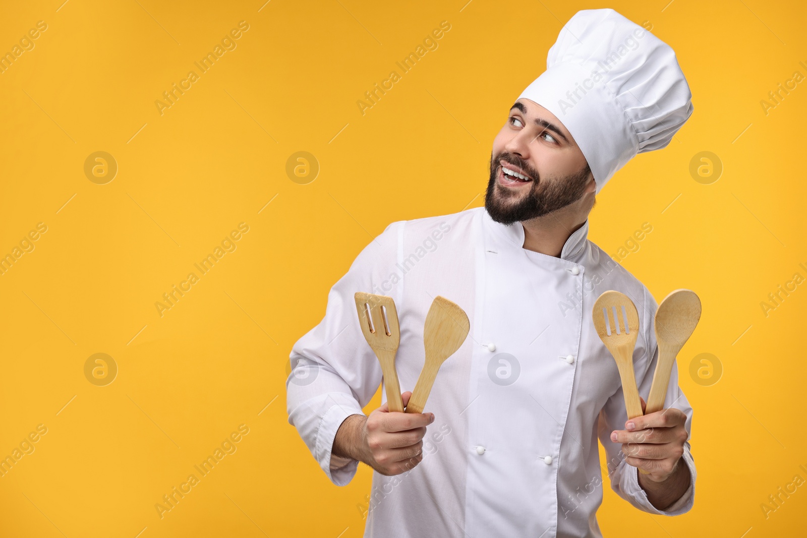 Photo of Happy young chef in uniform holding wooden utensils on orange background. Space for text