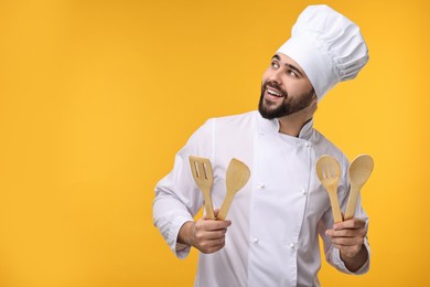 Photo of Happy young chef in uniform holding wooden utensils on orange background. Space for text