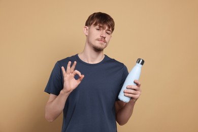 Man holding thermo bottle with drink and showing OK gesture on beige background