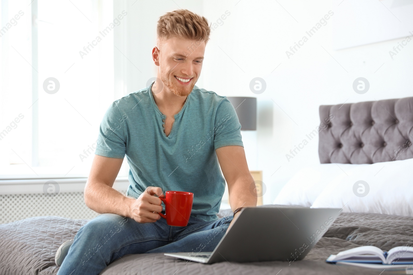 Photo of Young man using laptop while sitting on bed at home