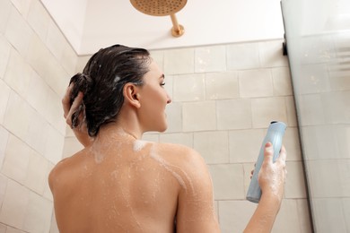 Woman with bottle of shampoo in shower at home, back view. Washing hair