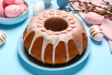 Photo of Delicious Easter cake decorated with sprinkles near painted eggs and willow branches on light blue background, closeup
