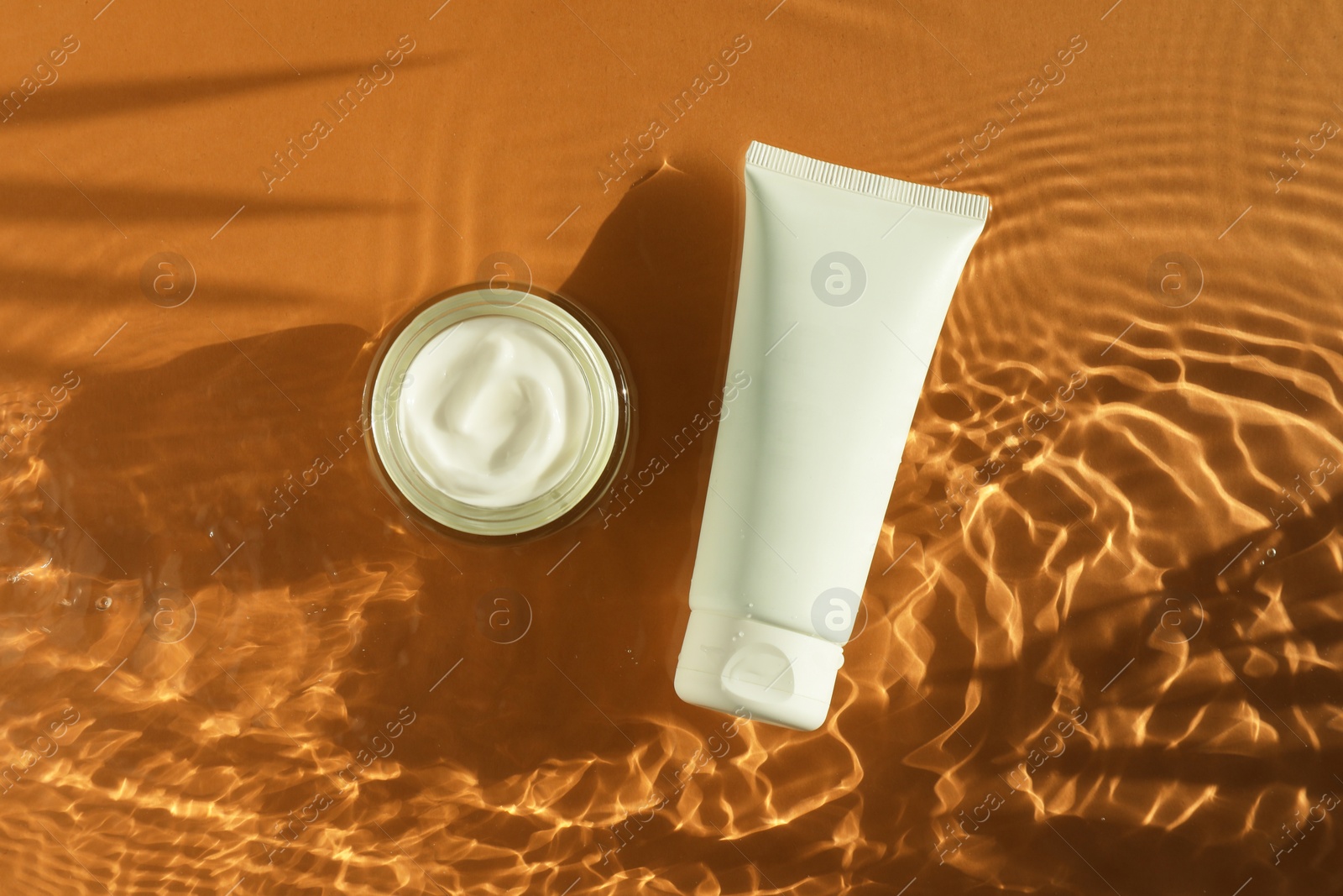 Photo of Tube and jar with moisturizing cream in water on orange background, top view