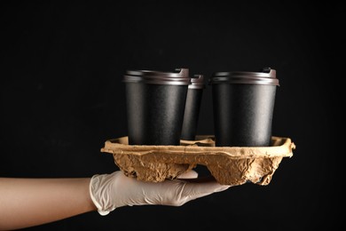 Woman holding cardboard holder with takeaway paper coffee cups on black background, closeup