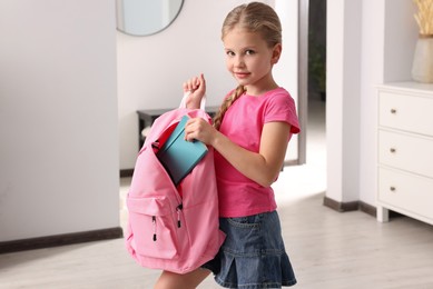 Photo of Little girl putting notebook in pink backpack at home. Preparing to school