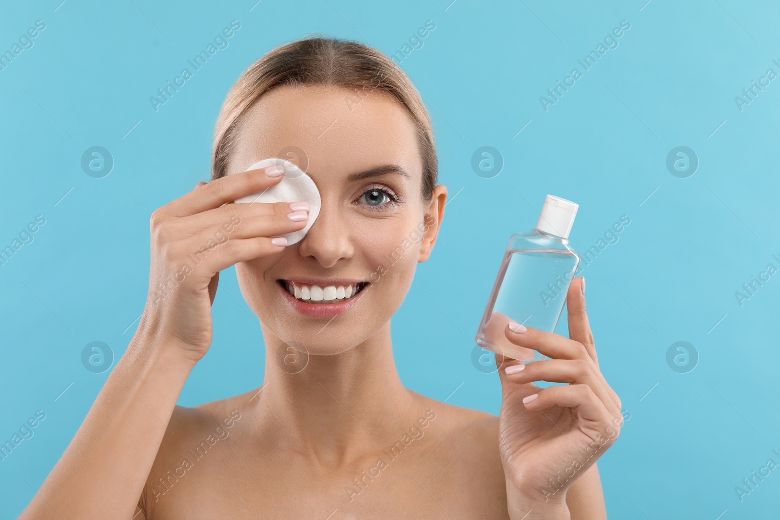 Photo of Smiling woman removing makeup with cotton pad and holding bottle on light blue background