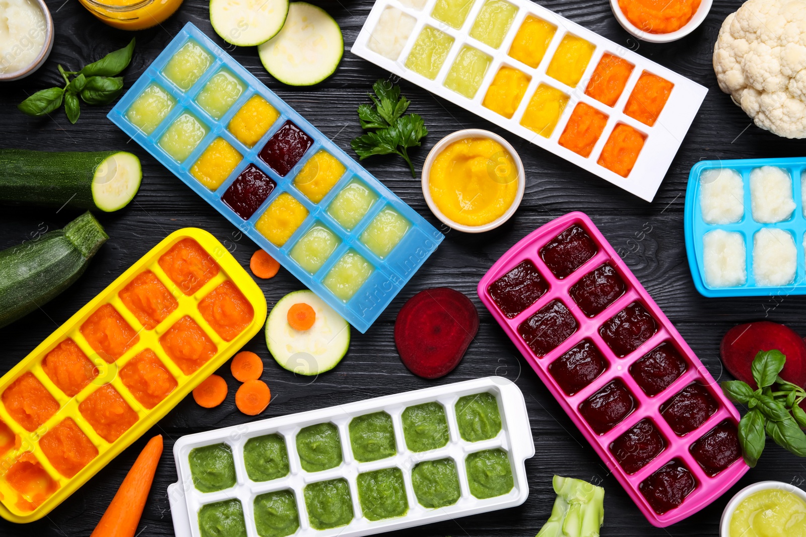 Photo of Different frozen purees in ice cube trays and ingredients of black wooden table, flat lay. Ready for freezing