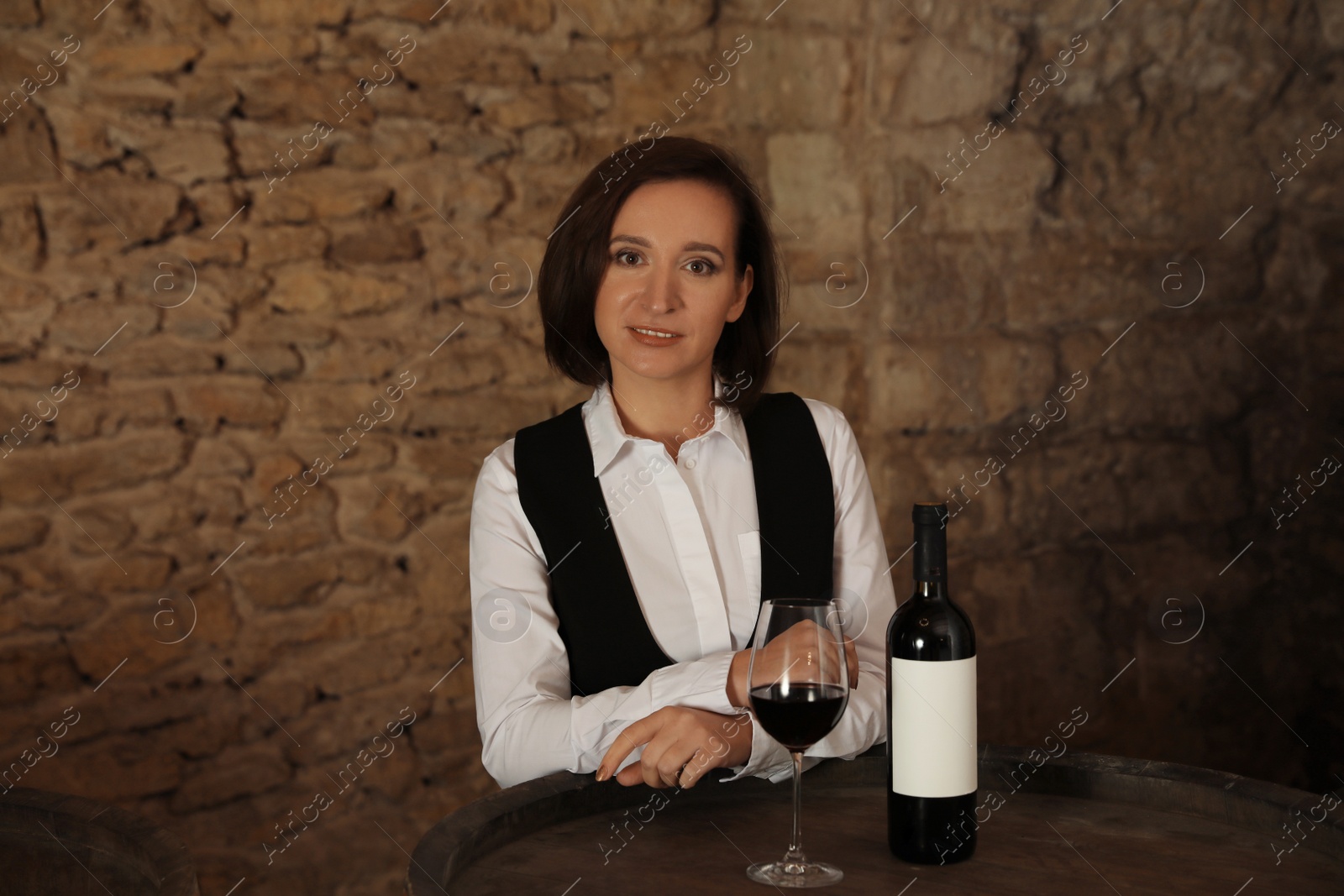 Photo of Female sommelier with glass and bottle of red wine at table indoors