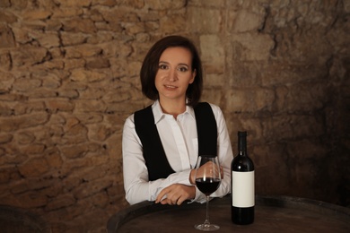 Photo of Female sommelier with glass and bottle of red wine at table indoors