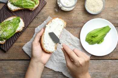 Photo of Woman spreading cream cheese onto bread for avocado sandwich at wooden table, top view