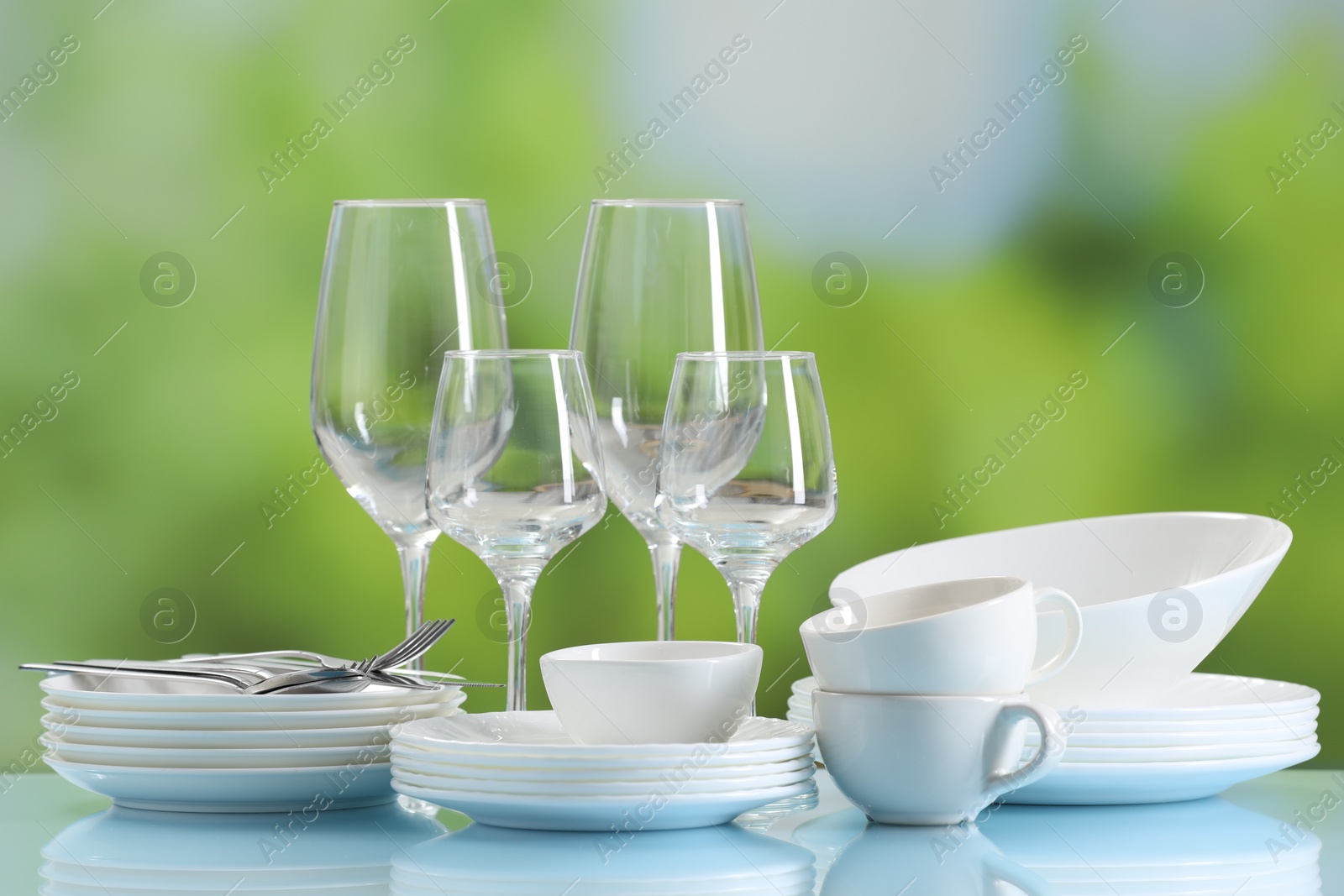 Photo of Set of many clean dishware, cutlery and glasses on light blue table against blurred green background