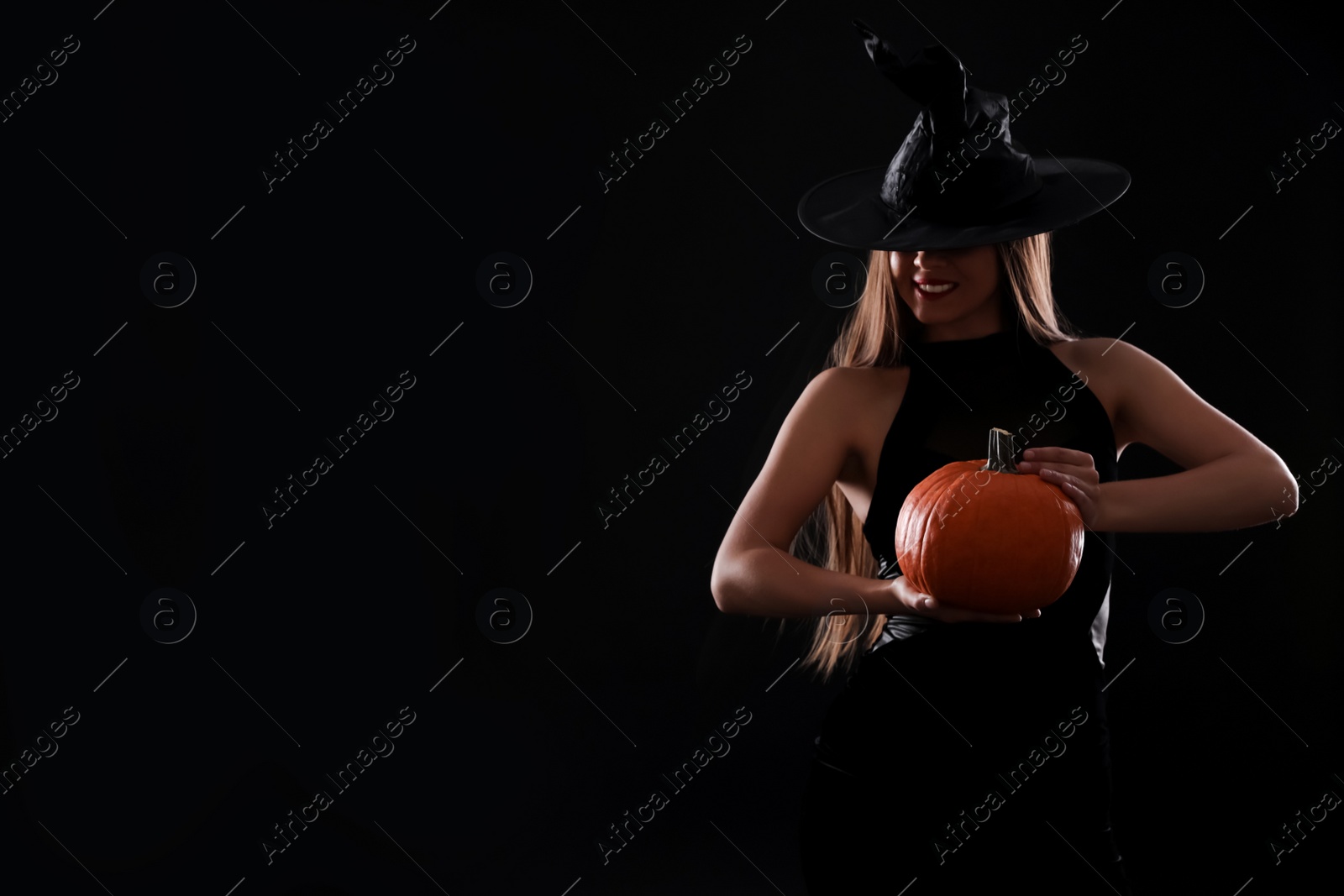Photo of Young woman wearing witch costume with pumpkin on black background, space for text. Halloween party