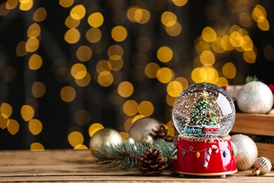 Beautiful snow globe and Christmas decor on wooden table against blurred festive lights. Space for text