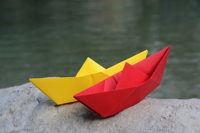 Photo of Beautiful yellow and red paper boats on stone near water outdoors, closeup