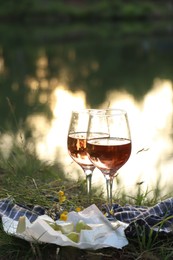 Photo of Glasses of delicious rose wine, cheese and grapes on picnic blanket near lake