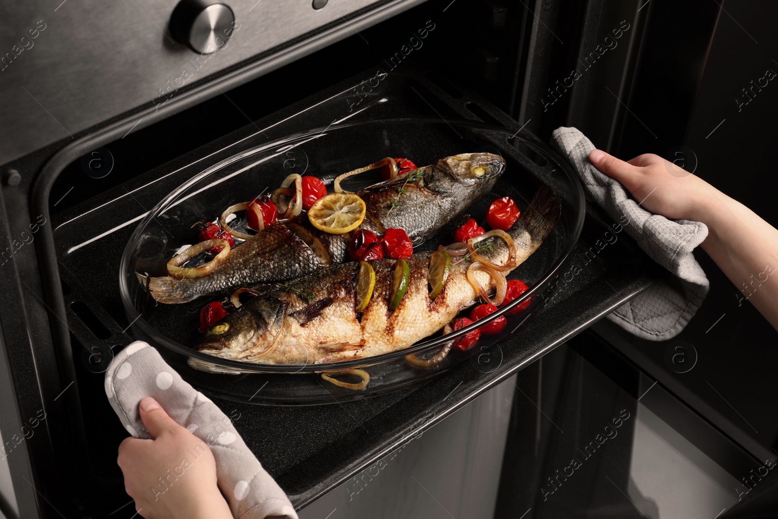 Photo of Woman taking out baking tray with sea bass fish and garnish from oven, closeup