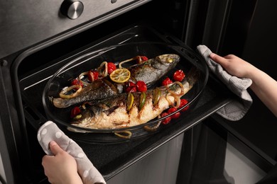 Woman taking out baking tray with sea bass fish and garnish from oven, closeup