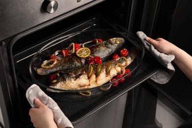 Photo of Woman taking out baking tray with sea bass fish and garnish from oven, closeup