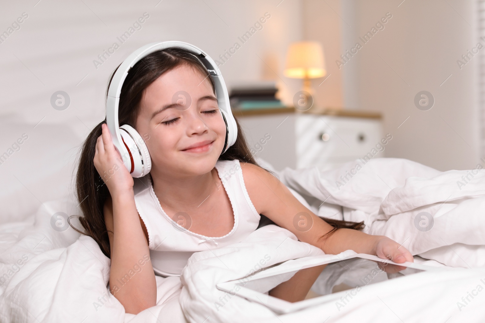 Photo of Cute little girl with headphones and tablet listening to audiobook in bed at home