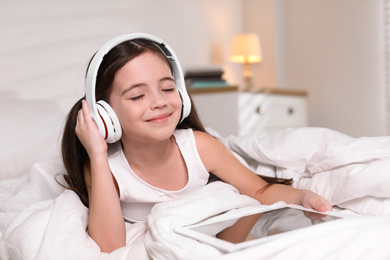 Photo of Cute little girl with headphones and tablet listening to audiobook in bed at home