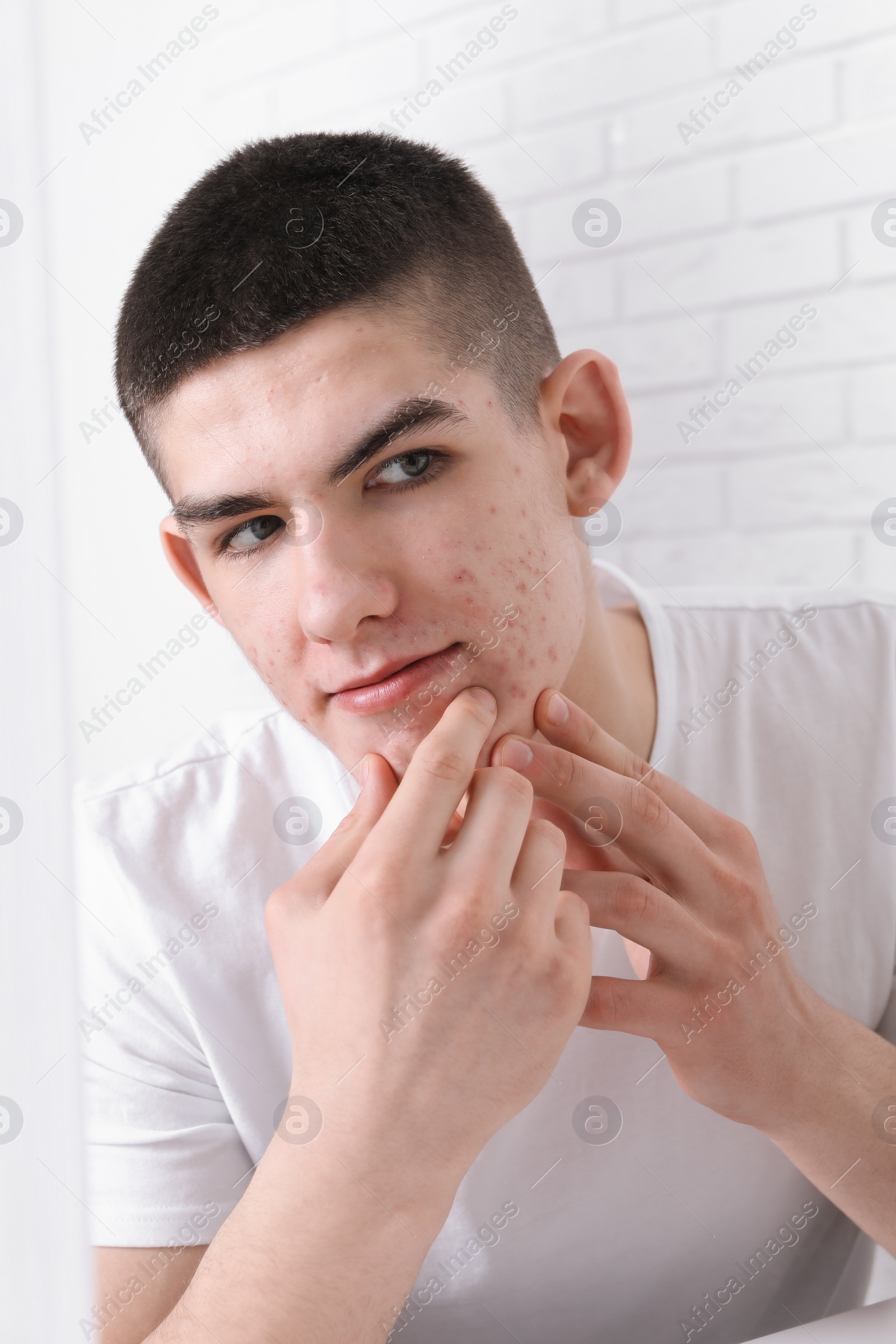 Photo of Young man looking at mirror and touching pimple on his face indoors. Acne problem