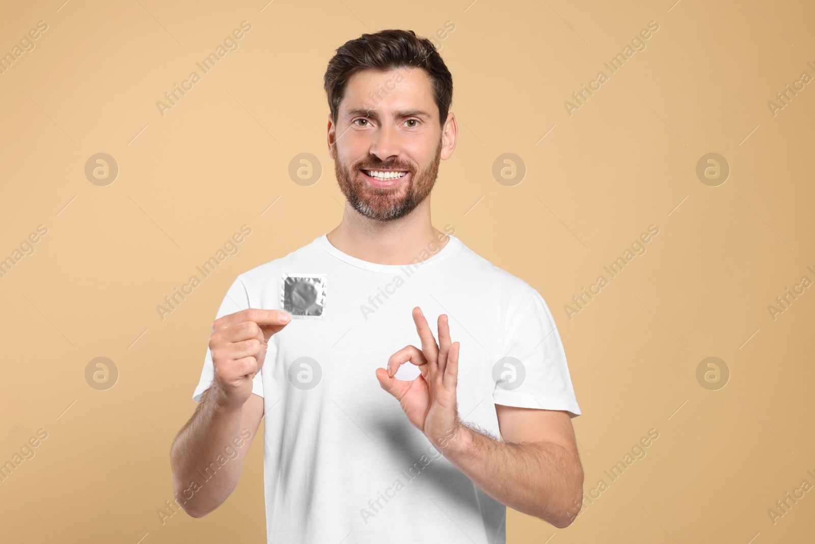 Photo of Man with condom showing ok gesture on beige background. Safe sex