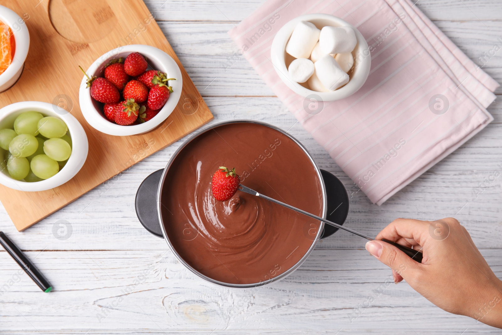 Photo of Woman dipping strawberry into pot with chocolate fondue on wooden background, flat lay