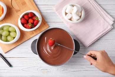 Photo of Woman dipping strawberry into pot with chocolate fondue on wooden background, flat lay