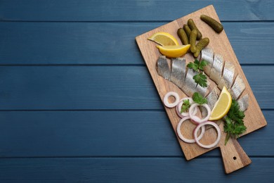 Sliced salted herring fillet served with parsley, pickles, onion rings and lemon on blue wooden table, top view. Space for text