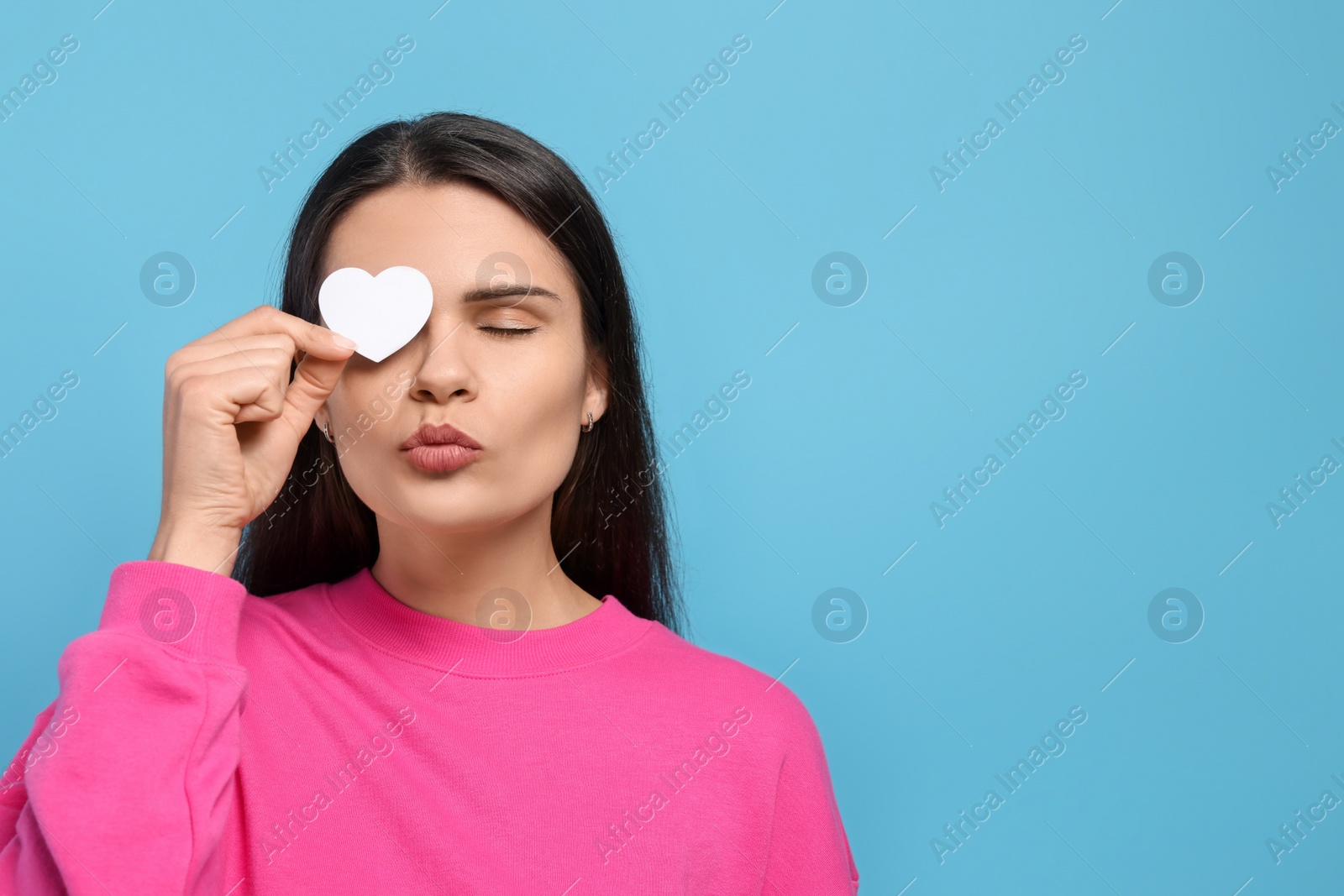 Photo of Beautiful young woman with paper heart giving kiss on light blue background. Space for text