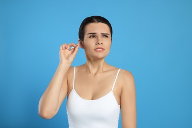Young woman cleaning ear with cotton swab on light blue background