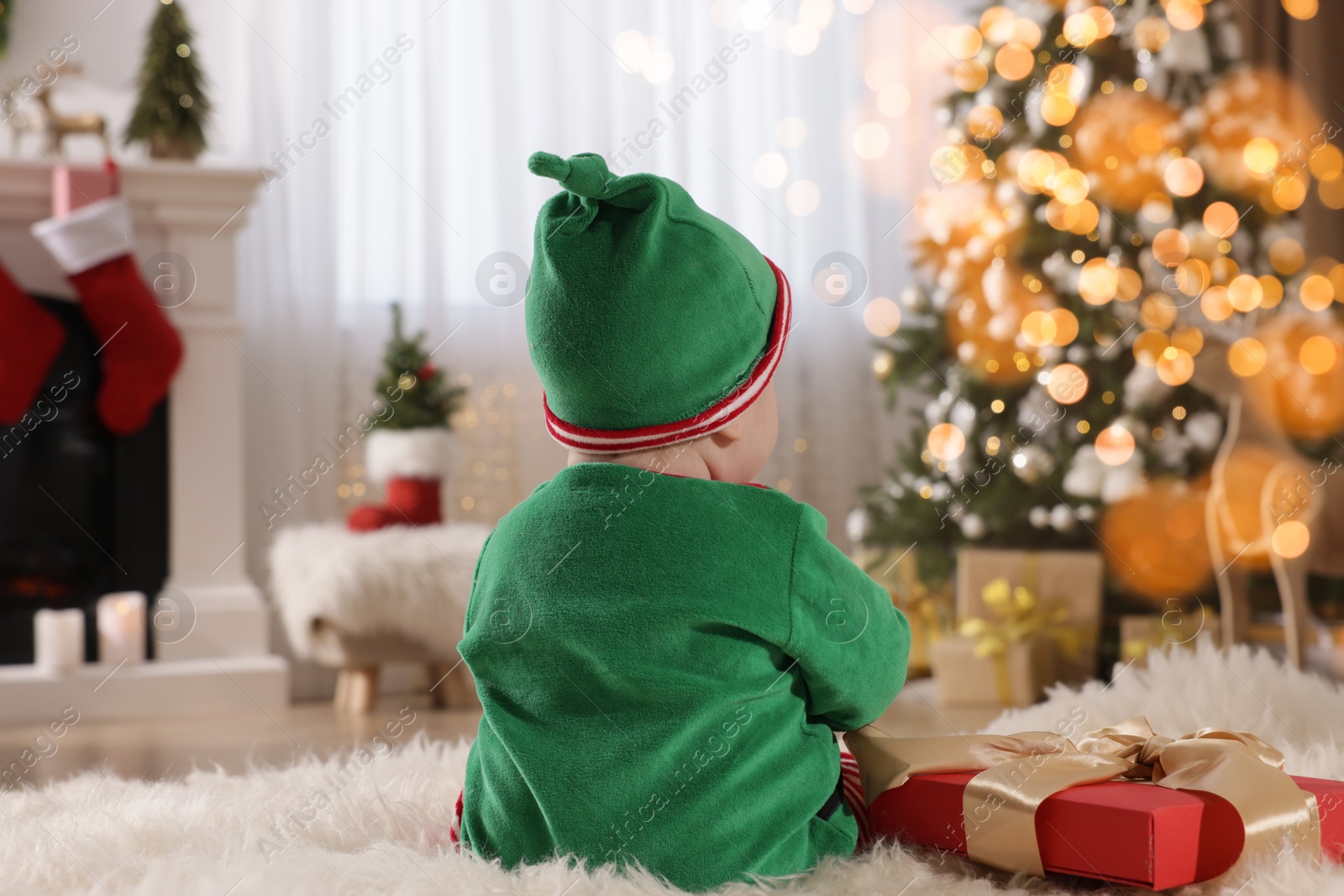 Photo of Baby in cute elf costume near Christmas gift on floor at home, back view