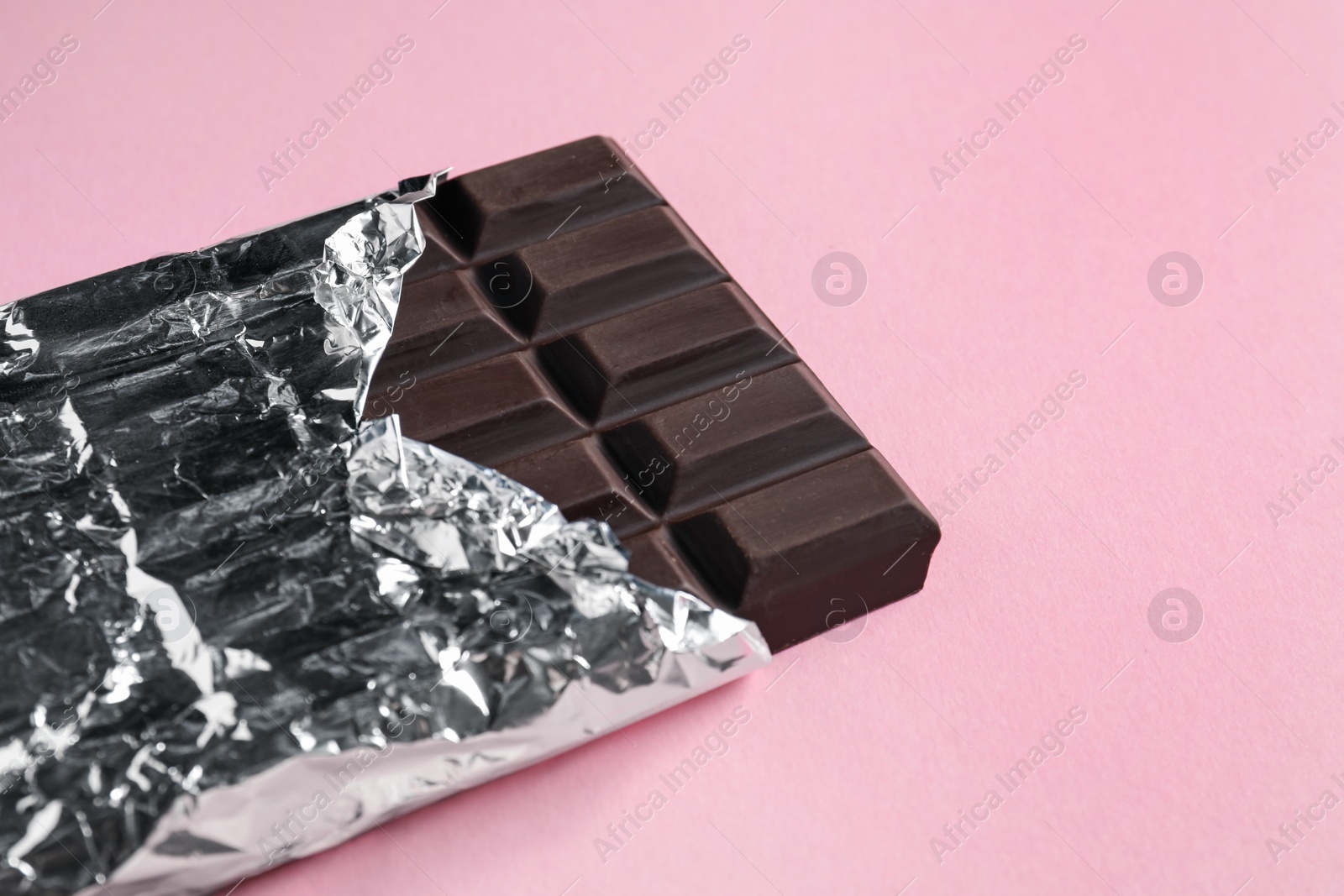 Photo of Delicious dark chocolate bar wrapped in foil on pink background. Space for text