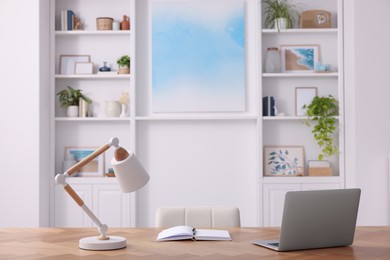 Photo of Interior design. Cosy workplace with laptop and lamp on wooden table near abstract picture between shelves