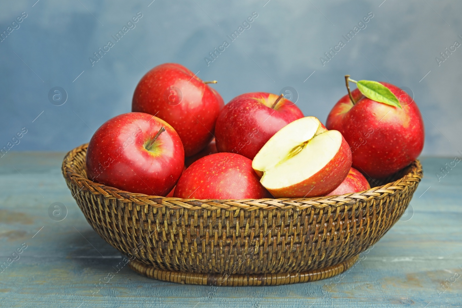 Photo of Wicker bowl with ripe juicy red apples on wooden table against blue background